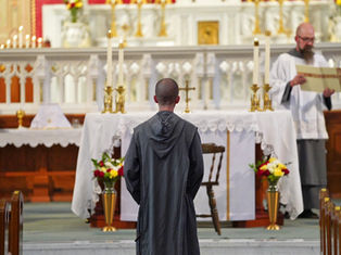 A monk in a gray robe stands before an altar in a church, with priests nearby carrying a scroll. The altar is adorned with candles and flowers.