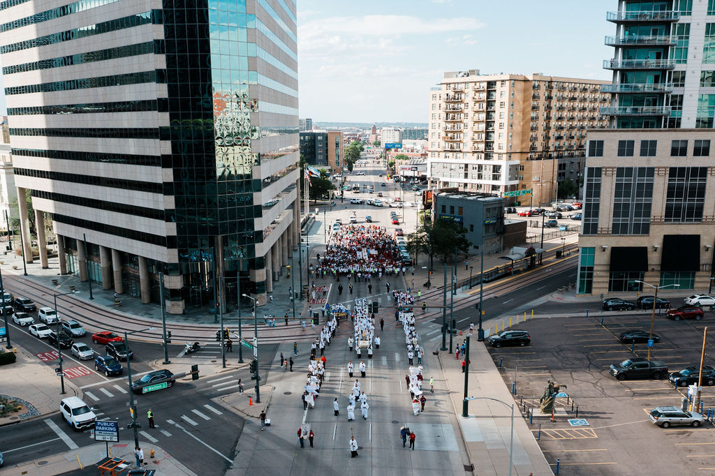 Large march with a crowd filling a downtown street between tall buildings under a bright sky.