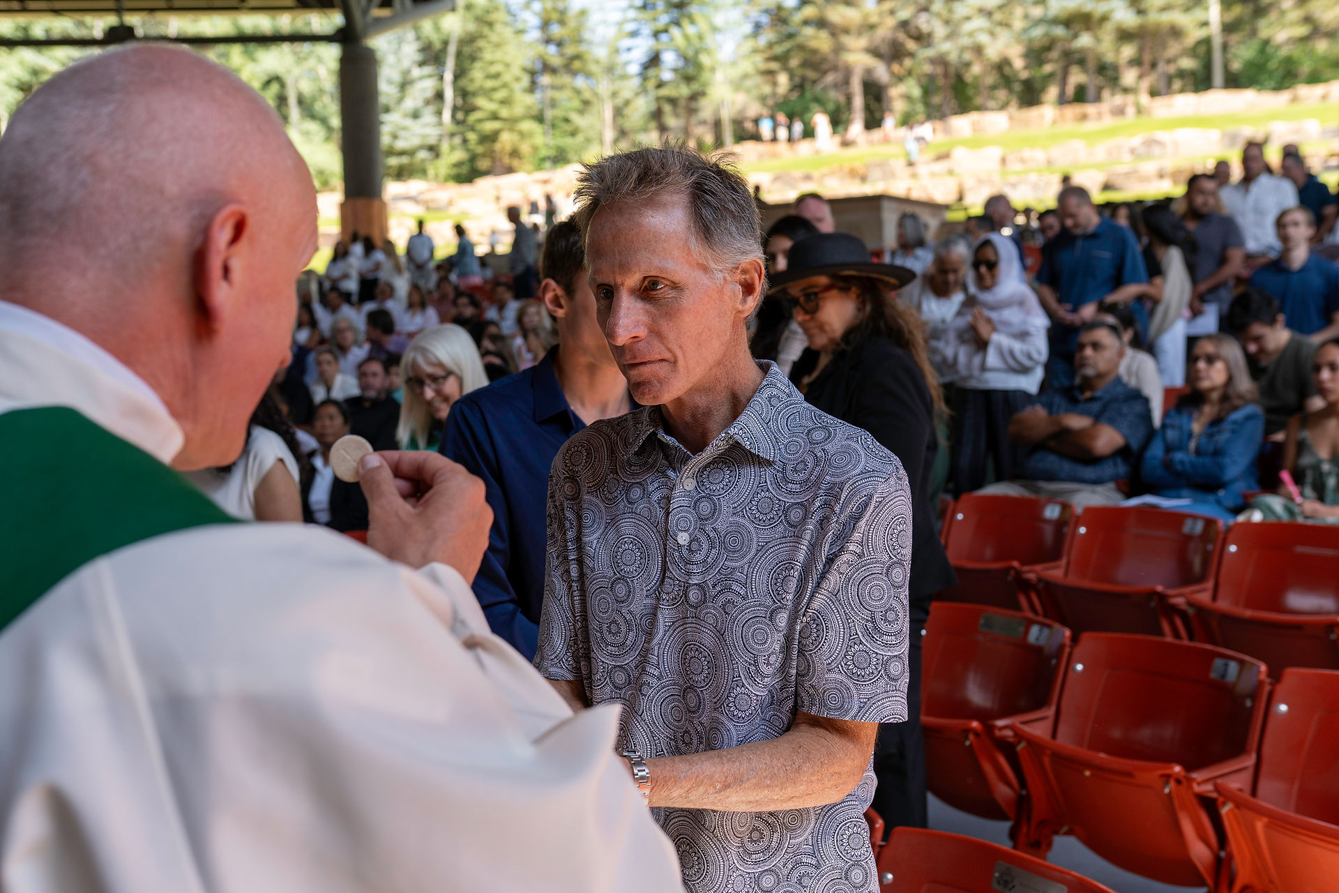 A person receives communion from a priest during an outdoor religious ceremony. Attendees are seated in red chairs and standing in the background, with trees and greenery surrounding the setting.