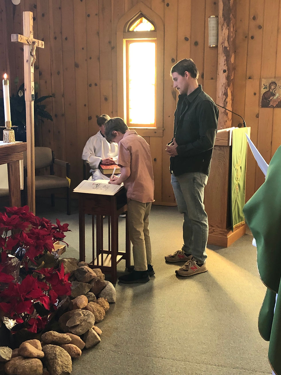 Boy writes in a church setting, accompanied by a man. A candle and red flowers adorn the wooden interior, with a sunlit window nearby.