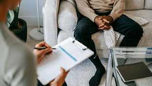 Person sits on a sofa with hands clasped; another takes notes on paper. Neutral tones and modern decor set a calming mood.