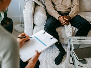 Person sits on a sofa with hands clasped; another takes notes on paper. Neutral tones and modern decor set a calming mood.