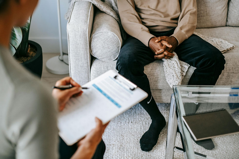 Person sits on a sofa with hands clasped; another takes notes on paper. Neutral tones and modern decor set a calming mood.