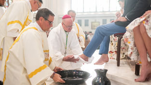 Clergy in white robes and red caps perform foot washing ceremony in a bright church, with a bowl and jug on the floor.