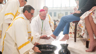 Clergy in white robes and red caps perform foot washing ceremony in a bright church, with a bowl and jug on the floor.