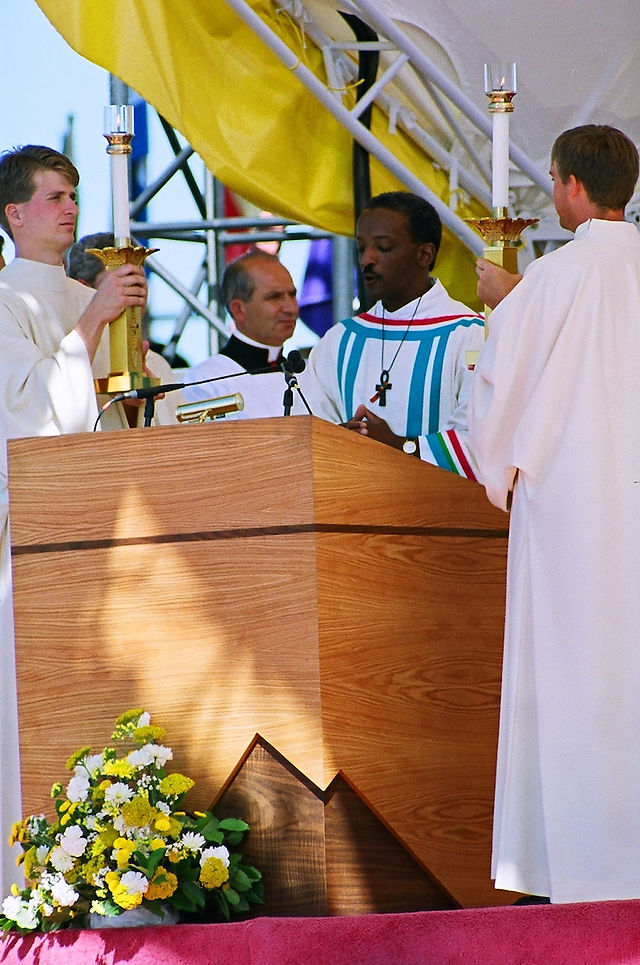 Altar servers at a wooden podium hold lit candles during a service. A man in a multicolored robe speaks. Yellow flowers adorn the podium.