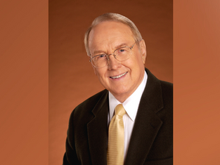 Smiling man in glasses wearing a dark suit and yellow tie against a warm brown background. Professional and welcoming mood.