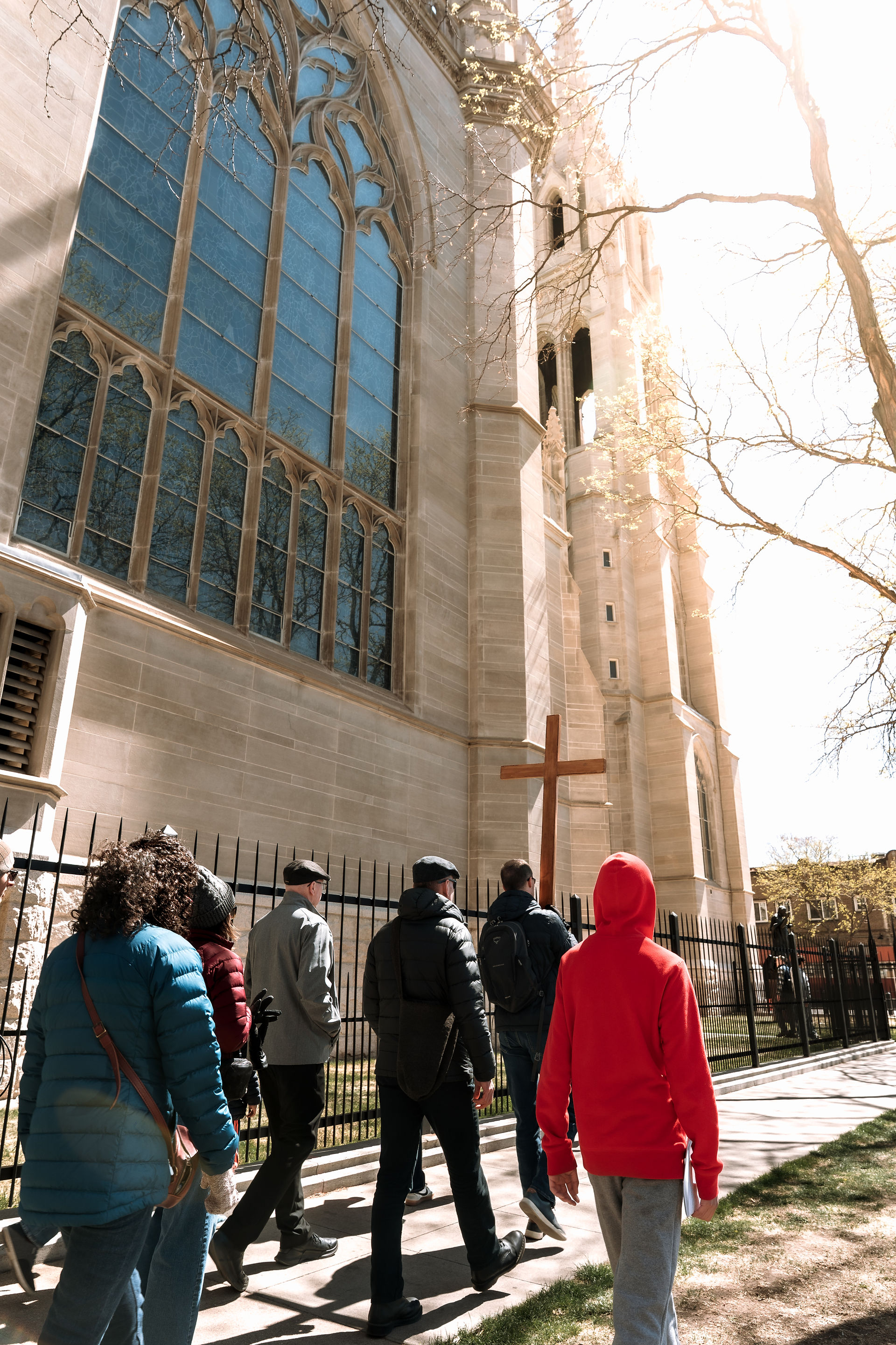 A group walks alongside a church building with stained glass windows, following a wooden cross during a prayer procession.