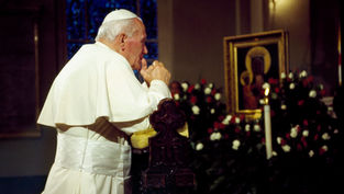 Man in white robe praying at a church altar, hands clasped. Icon and flowers in background, warm lighting, contemplative mood.