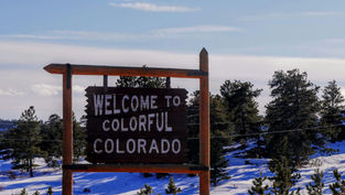 Wooden sign reading "Welcome to Colorful Colorado" amidst snowy trees under a clear blue sky.