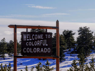 Wooden sign reading "Welcome to Colorful Colorado" amidst snowy trees under a clear blue sky.