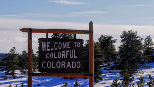 Wooden sign reading "Welcome to Colorful Colorado" amidst snowy trees under a clear blue sky.