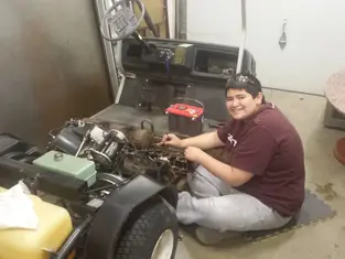 Teen fixing golf cart engine, smiling, in garage. Red toolkit on seat, assorted wires visible. Warm lighting, relaxed mood.
