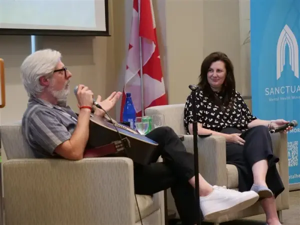 Man with guitar talks to woman seated on stage. Canadian flag and "Sanctuary" banner visible. Relaxed setting, casual conversation.
