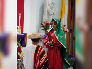 Children in traditional attire perform in a church. Girl in green and red, boy in sombrero. Candles, flowers, and statue in the background.