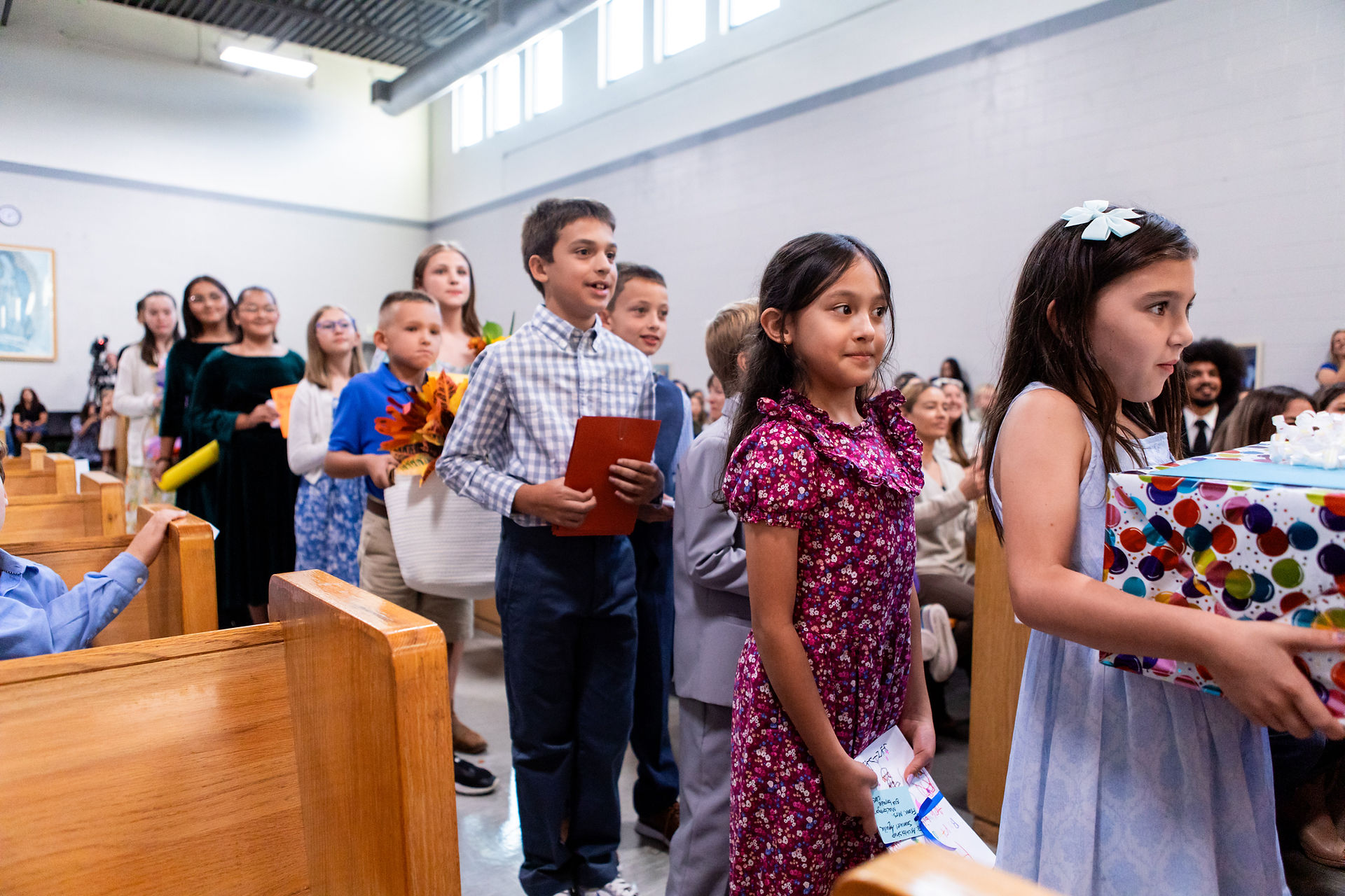 Children in formal attire line up in a church, one holding a colorful gift. They appear focused, with seated adults in the background.