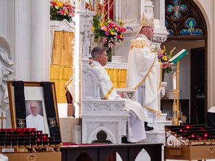 Priest in white robe speaks at pulpit in ornate church. Seated clergyman listens. Flowers and a framed photo decorate the setting.