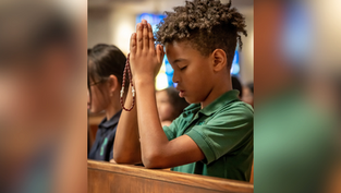 Boy in a green shirt praying with a rosary in church, eyes closed. Blurred stained glass in background, serene atmosphere.