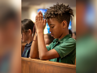 Boy in a green shirt praying with a rosary in church, eyes closed. Blurred stained glass in background, serene atmosphere.