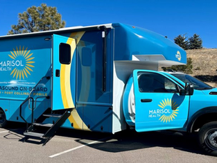 Blue mobile medical van labeled "Marisol Health" with ultrasound, parked outdoors on a sunny day. Stairs lead to an open side door.
