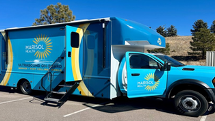 Blue mobile medical van labeled "Marisol Health" with ultrasound, parked outdoors on a sunny day. Stairs lead to an open side door.