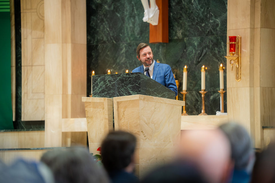 Man in a blue suit speaks at a church lectern before lit candles, with a blurred congregation in the foreground.