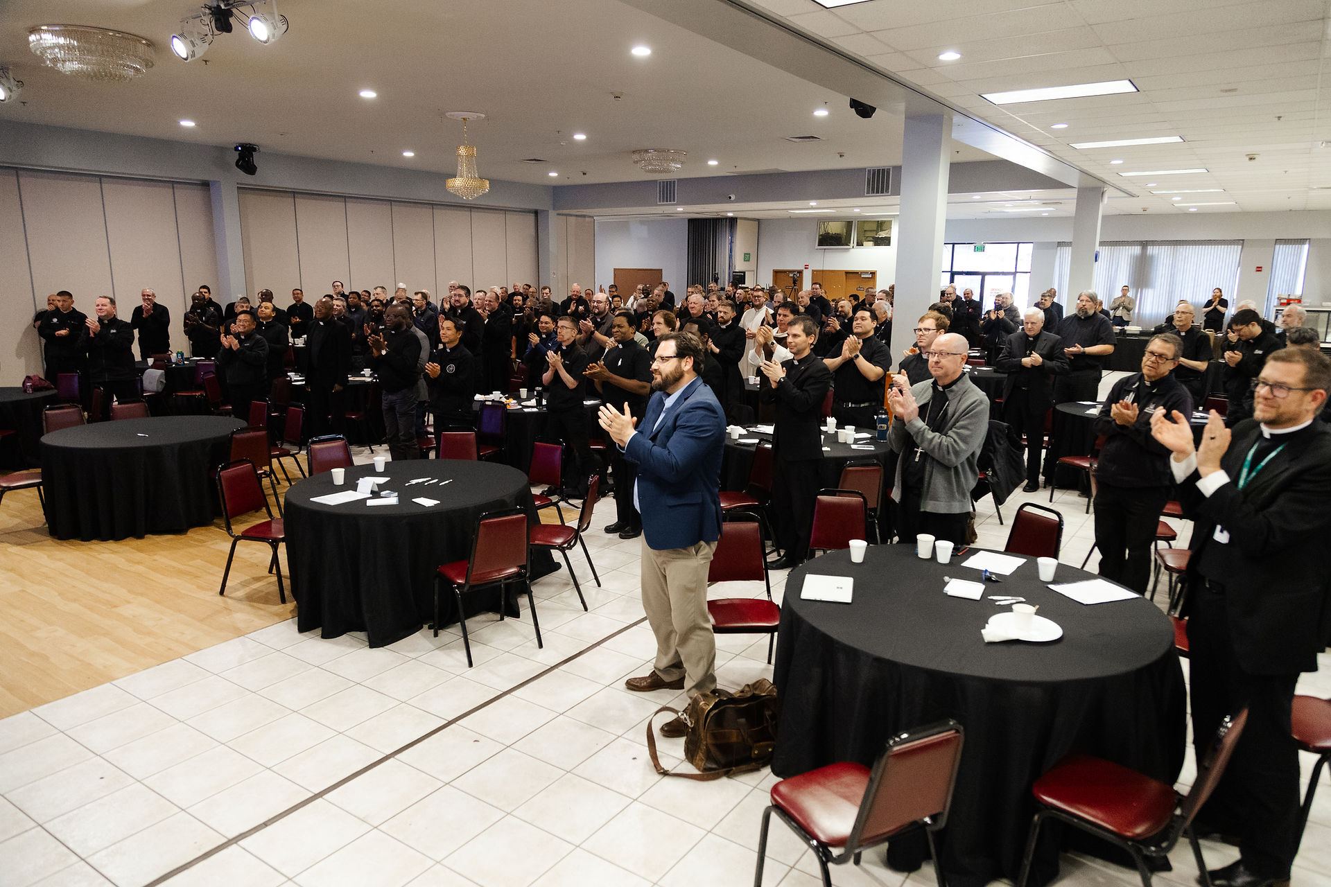 A large group of people stands and applauds at a conference in a banquet hall with black tablecloths and red chairs, creating an energetic atmosphere.