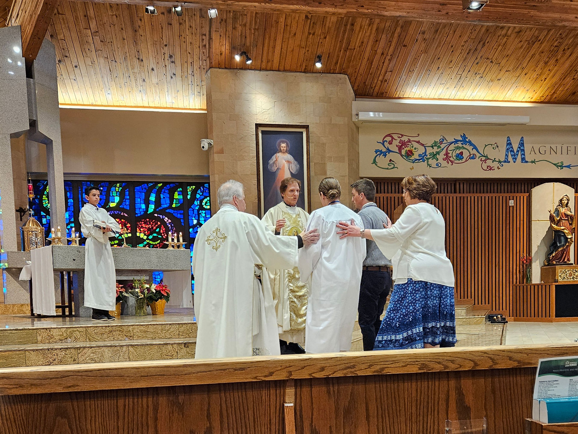 Priests and parishioners gather in a church with stained glass and wooden ceiling, creating a solemn, spiritual atmosphere.