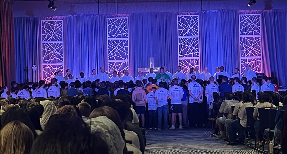 A group of people in white shirts face a stage with clergymen in robes. Blue and purple lighting creates a solemn ambiance, with decorative panels behind them.