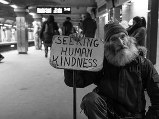 Bearded man in a beanie holds a sign reading "Seeking Human Kindness" in a subway station. People walk by in the background. Black and white.
