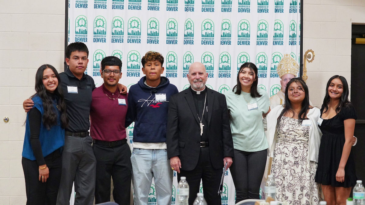 A group of eight people smiling, standing in front of an "Archdiocese of Denver" backdrop. Bottles on table in the foreground.