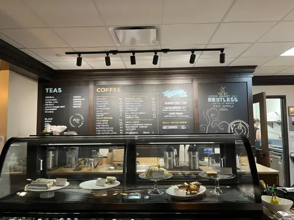 Cafe counter with desserts on display. Menus for teas and coffees on blackboard behind. Warm lighting with a cozy ambiance.