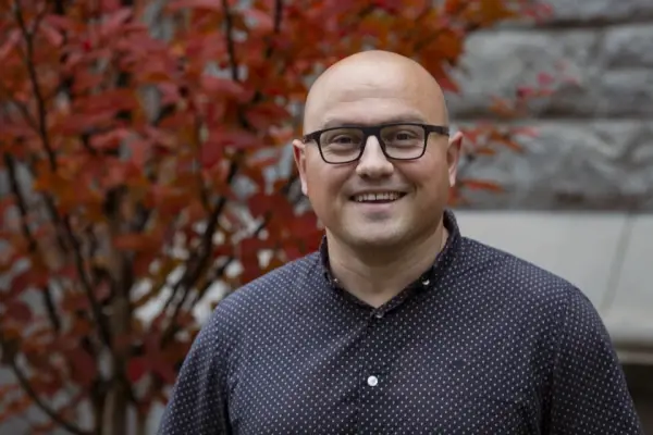 Smiling person with glasses in a dark polka dot shirt stands outdoors. Red leaves and a stone wall form the colorful autumn background.