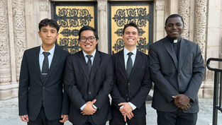Four men in black suits stand smiling in front of an ornate building with detailed stonework and decorative doors. The mood is formal and cheerful.