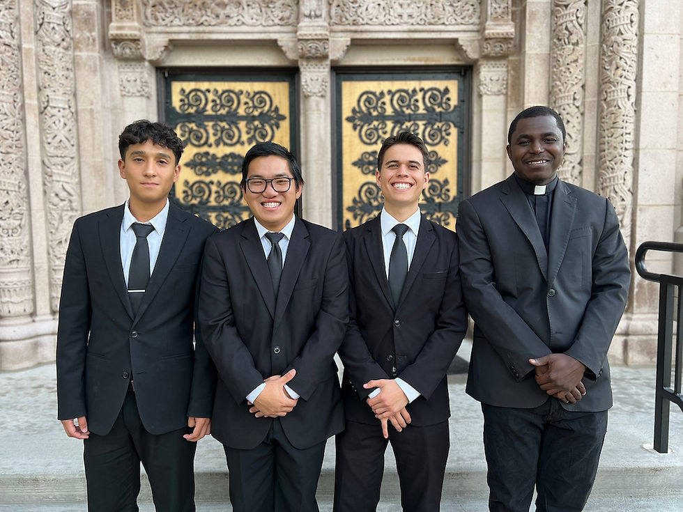 Four men in black suits stand smiling in front of an ornate building with detailed stonework and decorative doors. The mood is formal and cheerful.