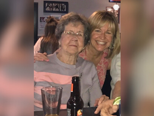 Two women smiling at a table with drinks; one wears glasses and gray striped top, the other a pink floral shirt. Warm, relaxed atmosphere.