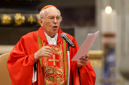 Elderly man in red religious robe speaks passionately into a microphone, holding paper. Candle and ornate background create solemn ambiance.