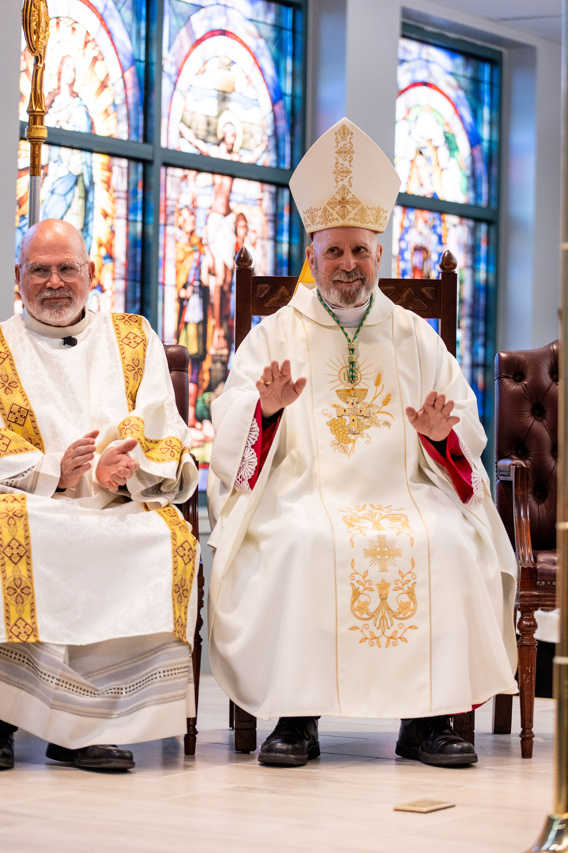 Two clergy members in ornate robes sit smiling, one claps. Stained glass windows in the background create a serene church ambiance.