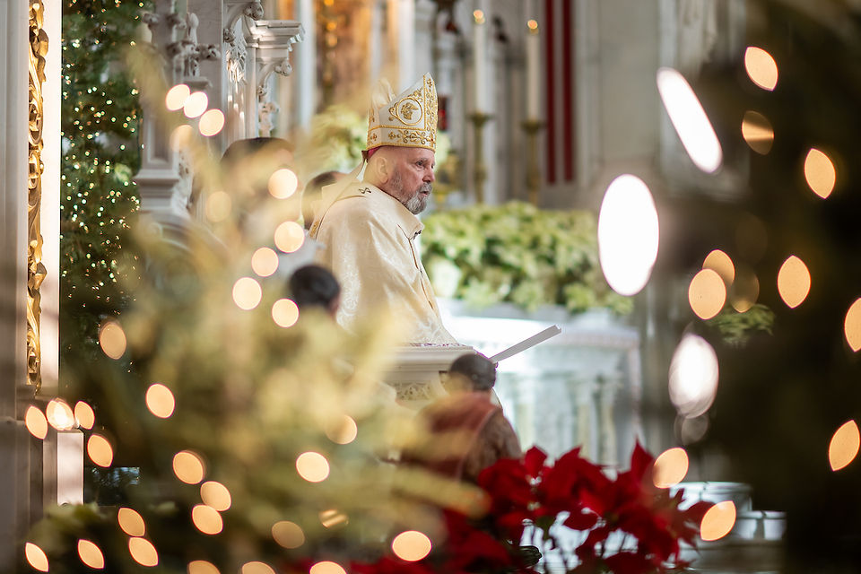 A bishop in ornate robes stands in a decorated church with Christmas lights, greenery, and red poinsettias, conveying a festive, solemn mood.