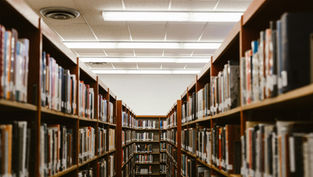 Library aisle with wooden bookshelves filled with books on both sides. Bright fluorescent lighting and a calm, quiet atmosphere.