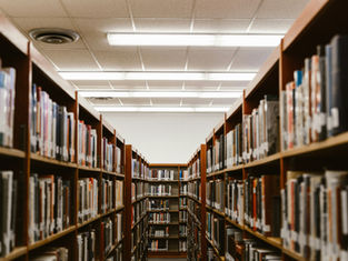 Library aisle with wooden bookshelves filled with books on both sides. Bright fluorescent lighting and a calm, quiet atmosphere.