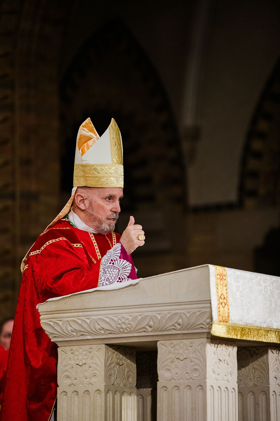 Bishop wearing red vestments and a gold mitre preaches from a stone ambo inside a church.