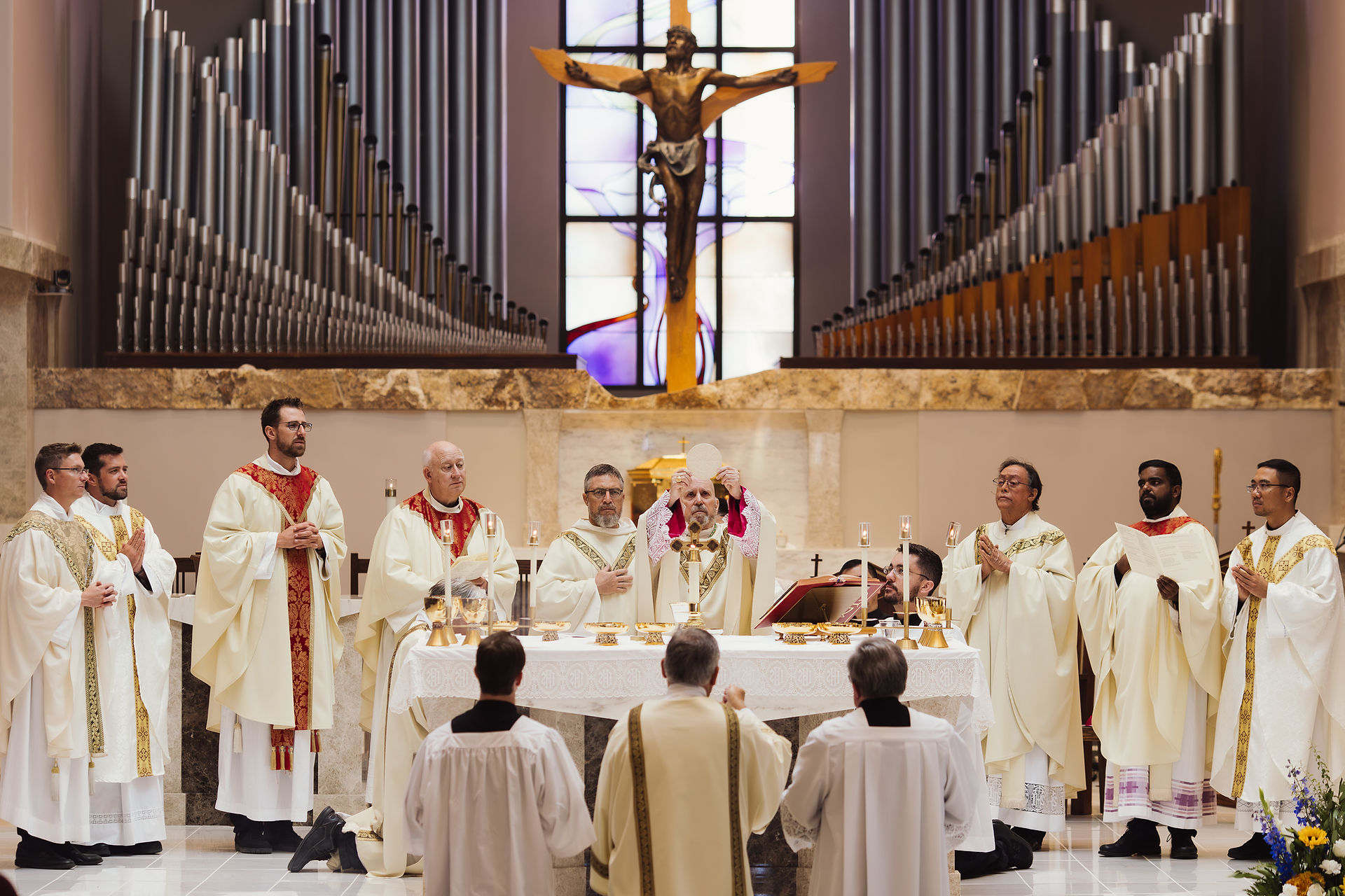 Priests in white and red vestments at an altar during a church service, one lifting a host. Stained glass and organ pipes in background.