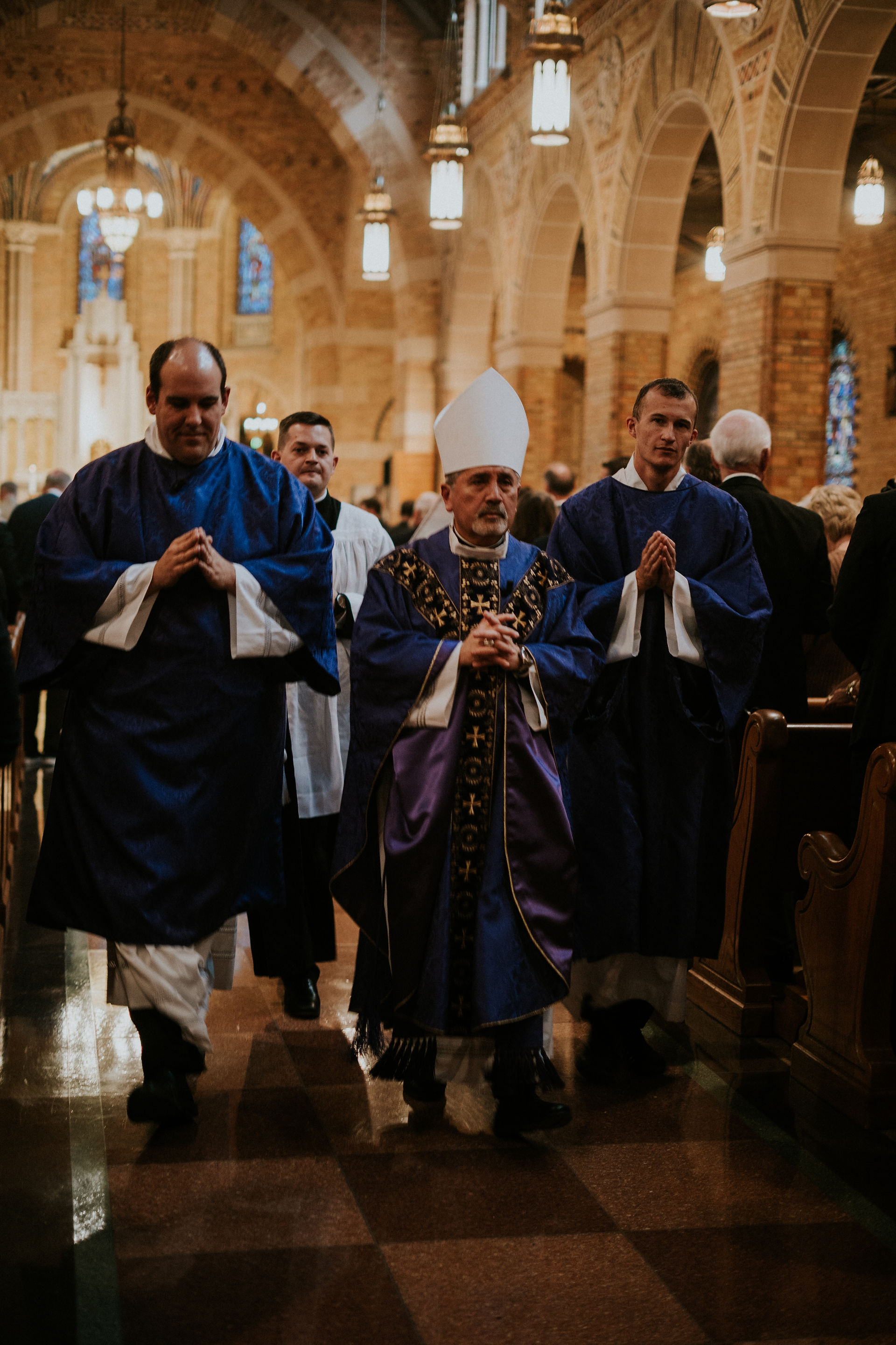 Clergy in purple robes walk down a church aisle. The setting is ornate with arched ceilings and chandeliers. Mood is solemn.
