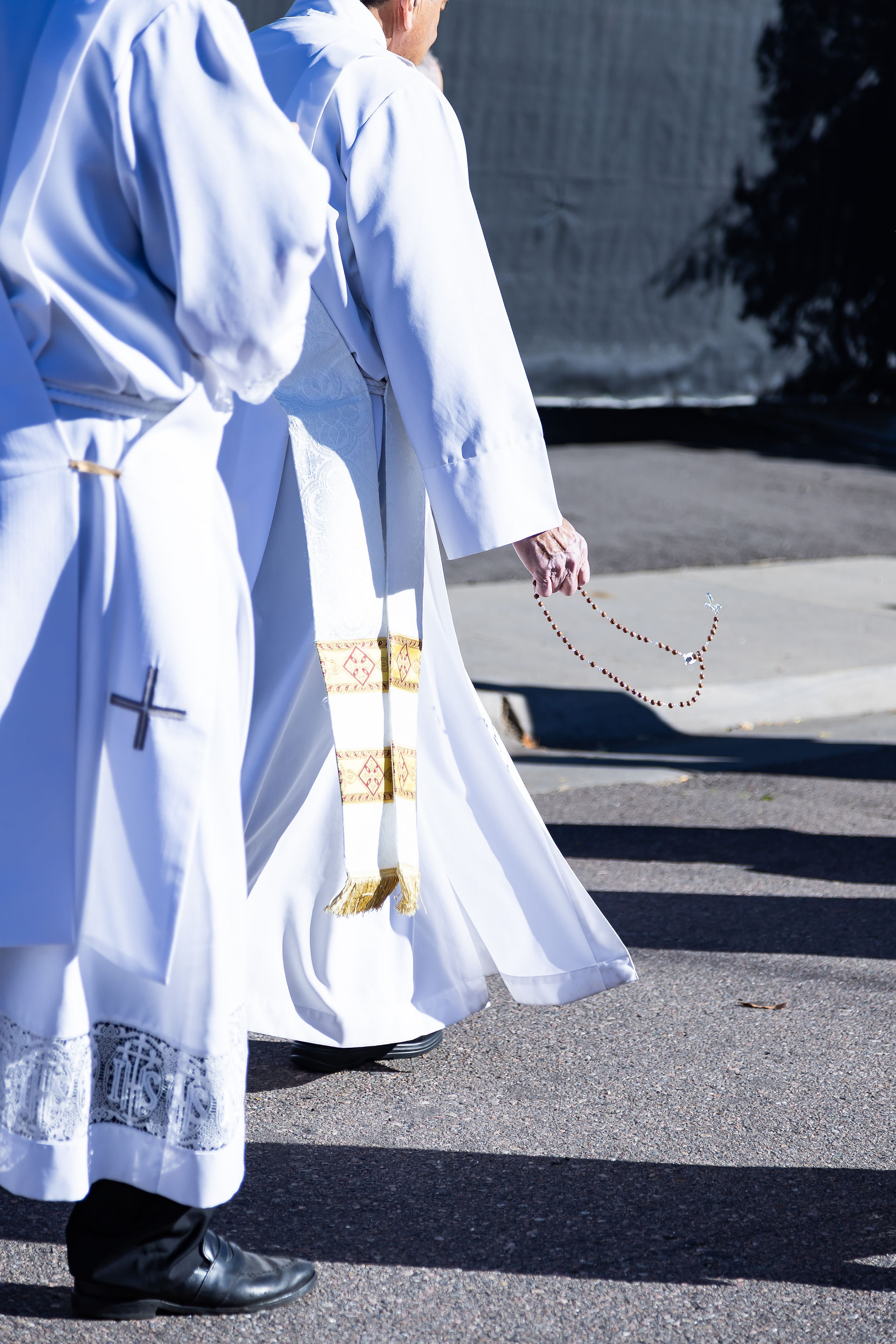 Clergy in white robes with sashes walk on a sunny pavement. One holds a rosary. Background is dark and shadowed. Peaceful mood.