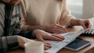 Two people, focused, review documents at a wooden table with a keyboard, coffee mug, and notebook in view. Natural light brightens the scene.