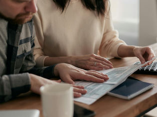 Two people, focused, review documents at a wooden table with a keyboard, coffee mug, and notebook in view. Natural light brightens the scene.