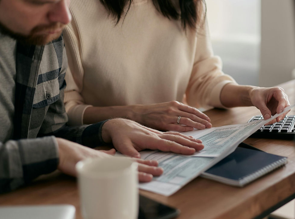 Two people, focused, review documents at a wooden table with a keyboard, coffee mug, and notebook in view. Natural light brightens the scene.