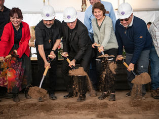 People in hard hats and colorful attire participate in a joyous groundbreaking ceremony, shoveling dirt indoors.