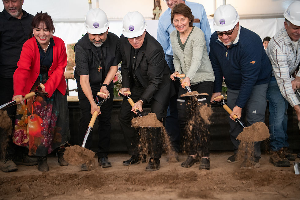 People in hard hats and colorful attire participate in a joyous groundbreaking ceremony, shoveling dirt indoors.